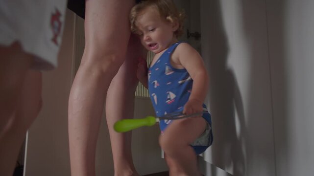 Toddler standing close to adult legs in kitchen holding green plastic step stool with cabinets and household items in background