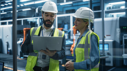 Hispanic Male Industrial  Engineer Using Laptop Computer And Talking To a Caucasian Female Designer At an Autonomous Assembly Line With Robotic Arms. Colleagues Monitoring Machinery Production.