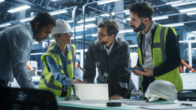 Meeting At Electronics Factory Office: Diverse Specialists Using Laptop And Tablet With Blueprints For Innovative Green Energy Engine. Facility With Autonomous Assembly Line With Robotic Arms.