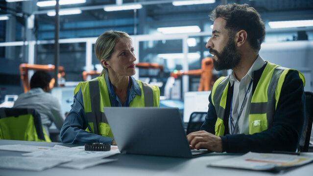 Factory Office: Caucasian Female Quality Control Technician Talks to Hispanic Male Industrial Engineer Working on Laptop Computer. Automated Robot Arm Assembly Line Manufacturing Modern Engines.