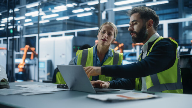 Autonomous Factory Office: Caucasian Female Quality Control Technician Talks to Hispanic Male Industrial Engineer Working on Laptop Computer. Automated Robot Arm Assembly Line Manufacturing Machinery.