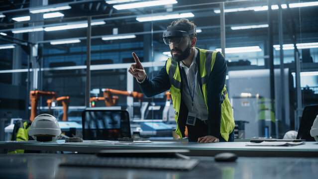 Hispanic Male Industrial Designer Sitting in Factory Office And Using Augmented Reality Headset To Adjust Autonomous Assembly Line With Robotic Arms. Man Gesturing, Using Futuristic Interface.