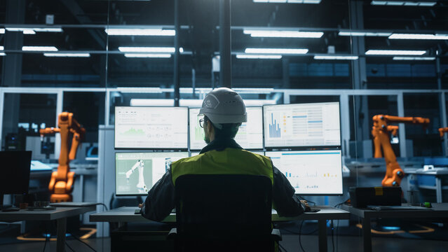 Backview of Caucasian Male Engineer Using Multi Monitor Workstation To Control Production Of Machinery on Autonomous Assembly Line With Robotic Arms - Powered by Adobe