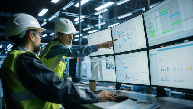 Caucasian Male Process Engineer Using Multi Monitor Workstation And Talking To Female Quality Control Technician At Electronics Factory. Colleagues Monitoring Autonomous Assembly Line With Robot Arms.
