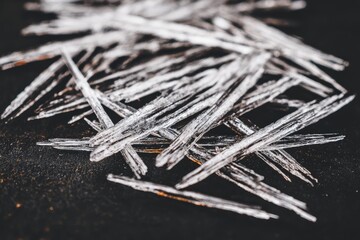 Close-up of many small, ice-like, sliver-shaped crystals on dark surface
