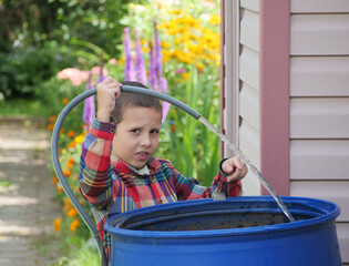 Young boy in a plaid shirt helps in the garden, filling a large blue rain barrel with water from a hose on a sunny August day