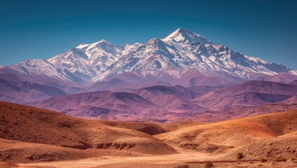 Majestic snow-capped mountain range dominates a vibrant desert landscape under a clear blue sky; rolling hills of reddish-brown contrast with purple-hued mountains in the mid-ground