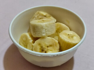 Ripe banana slices served in a white bowl on a plain pink cloth background.