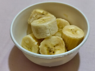Ripe banana slices served in a white bowl on a plain pink cloth background.