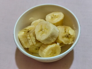 Ripe banana slices served in a white bowl on a plain pink cloth background.