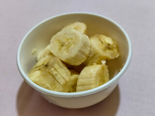 Ripe banana slices served in a white bowl on a plain pink cloth background.