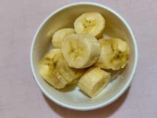 Ripe banana slices served in a white bowl on a plain pink cloth background.