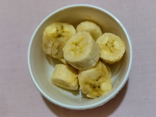 Ripe banana slices served in a white bowl on a plain pink cloth background.
