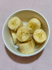 Ripe banana slices served in a white bowl on a plain pink cloth background.