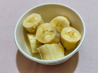 Ripe banana slices served in a white bowl on a plain pink cloth background.