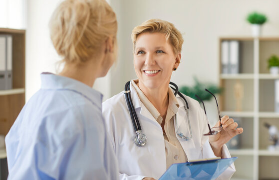 Smiling senior female doctor holding glasses and clipboard while attentively listening to young adult woman during medical consultation in modern clinic examination room. Healthcare services concept