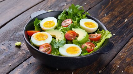 Close-up of a fresh healthy salad in a black bowl on a rustic wooden table. The dish includes soft-boiled eggs with runny yolks, sliced avocado, cherry tomatoes, lettuce, arugula, and sesame seeds. - Powered by Adobe