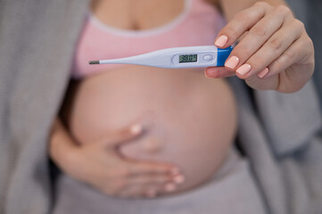 Pregnant woman measures temperature while sitting on the couch. Close up of the belly.