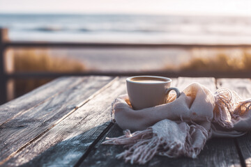 Warm cup of coffee on a beach table with scarf and soft golden light. Peaceful early autumn morning near the sea, perfect cozy moment -
