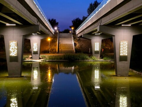 Bucharest, Romania - Illuminated pedestrian bridge in Tineretului Park at night, with glowing decorative panels mirrored perfectly in the still lake water.