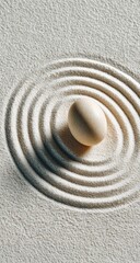 A single, off-white egg rests in the center of concentric circles raked into pale sand, creating a serene, minimalist scene