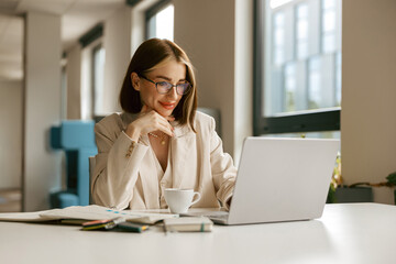 A very determined woman is seated in a stylish office, engrossed in her laptop while enjoying a...