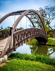 Wooden arched bridge over calm water, surrounded by lush green grass and flowering plants