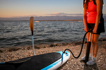 Sportswoman inflating paddle board with manual pump on a beach at sunset, preparing for paddle boarding