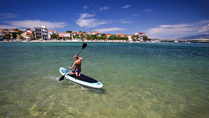 Young boy enjoying stand up paddleboarding in shallow turquoise water near a beach resort on a sunny summer day