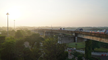Wide-angle view of a busy city bridge at sunrise, with vehicles crossing under soft morning light and lush green areas on either side, creating a dynamic urban scene