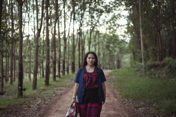 Young woman walking along a serene path in a lush green forest surrounded by tall trees, wearing a casual outfit and holding a bag, nature scene