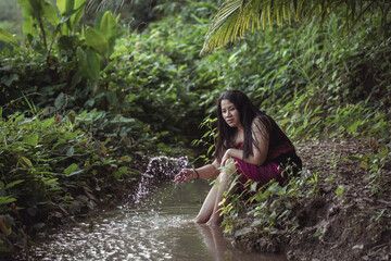 Fototapeta premium Serene Young Woman Sitting by a Stream in a Lush Green Forest, Enjoying Nature, Capturing the Essence of Tranquility and Peaceful Reflection