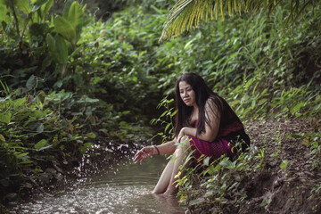 Fototapeta premium Young Woman Sitting by Tranquil Stream in Lush Green Forest, Enjoying Nature and Serenity, Reflecting on Life with Gentle Water Ripples