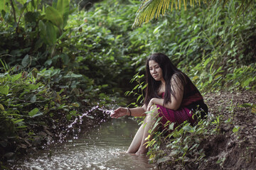 Fototapeta premium Young woman sitting by a serene stream in lush green foliage, gently splashing water while enjoying peaceful nature in a tropical setting