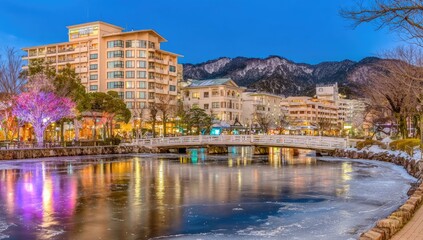 Panoramic view of a town by a frozen lake at twilight.  Colorful buildings, a bridge, and mountains in the background.  Illuminated trees and reflections on the ice-covered water