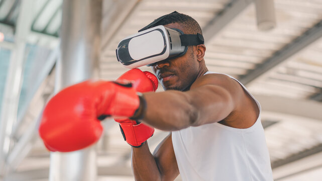 African-American man practices boxing with red gloves and VR headset under a modern urban shelter. Shows the integration of virtual reality, AI training, and tech-based fitness enhancement.