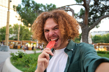 Fototapeta premium Ginger man taking a selfie while biting a popsicle in a park during summer