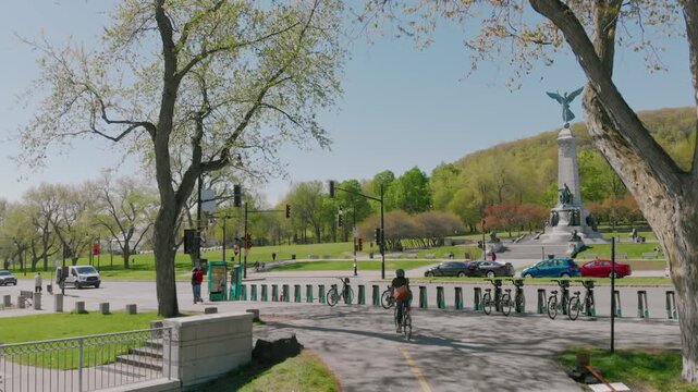 A low angle view of a road in Mont Royal, Montreal, Quebec, Canada, with bicycles parked near a monument. The road leads up a hill with trees and a cross in the background.