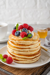 A stack of pancakes with whipped cream, fresh berries and honey on a white plate on a gray concrete background.