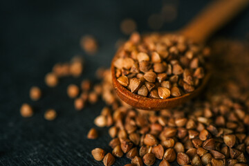 Close-up of raw buckwheat groats in a wooden spoon, scattered on a dark rustic surface....