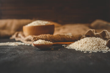 Top view of raw white rice in a wooden bowl, spoon, and burlap sack on a dark textured background. Rustic kitchen setup with natural food ingredients