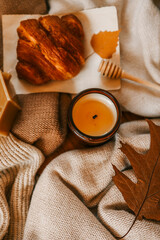 Cozy autumn scene with a golden croissant on parchment, honey dipper, lit candle in a brown glass jar, and a brown maple leaf. Beige knitted blankets create a warm, inviting background