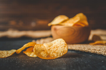 Crispy potato chips in a rustic wooden bowl on burlap fabric, with scattered chips on a dark...