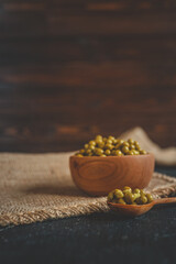 Close-up of green canned peas in a wooden bowl and spoon on a rustic fabric and dark background. Fresh, glossy texture, perfect for salads and side dishes