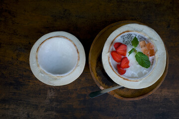 A mixed fruit salad, colorful dessert in half a fresh coconut, on a dark wooden table
