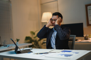 Stressed Asian businessman screaming and holding his head in his hands at his office desk