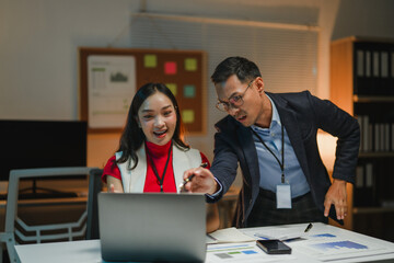 Asian businesspeople working late using laptop in office at night, surprised by something on screen
