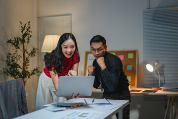 Asian business team celebrating success looking at laptop in office at night