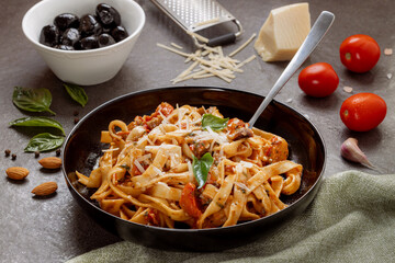 Italian tagliatelle pasta with sun-dried tomato sauce, parmesan cheese, fresh basil, and black olives served in a rustic bowl on stone table, surrounded by cherry tomatoes, garlic, and almonds, 