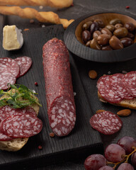 Gourmet Italian salami sausage sliced on black wooden serving board with artisan cheese, olives, grapes, almonds, and breadsticks, rustic stone background, daylight